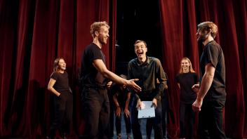 Eine Gruppe Menschen stehen gemeinsam vor einem roten Vorhang auf der Bühne und lachen.