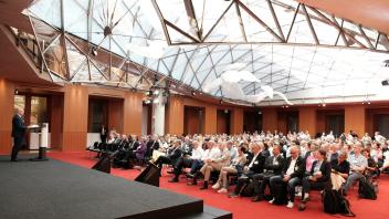 Publikum sitzt in einem großen Saal mit rotem Teppich und Glasdach und blickt auf eine Bühne