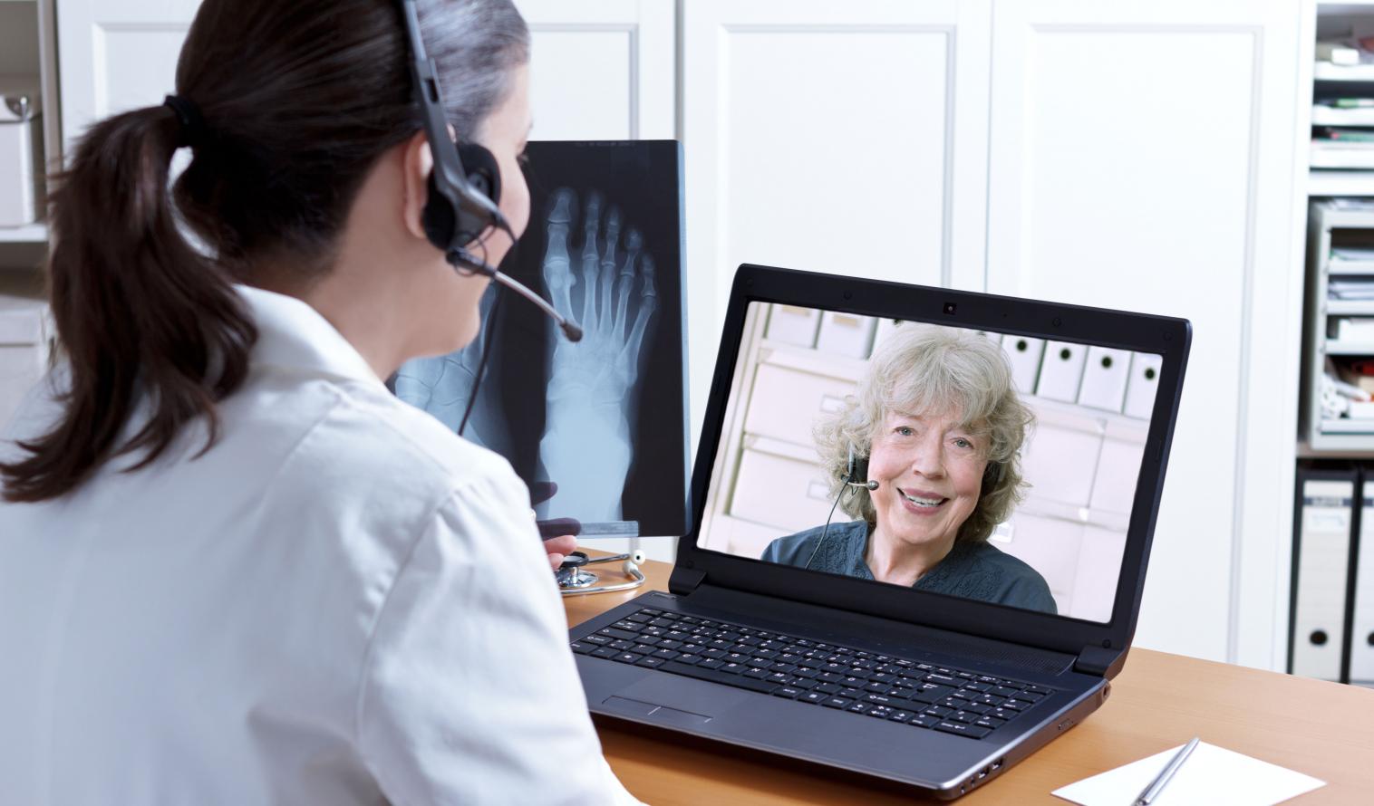 Ärztin in ihrem Sprechzimmer mit Headset vor ihrem Laptop, ein Röntgenbild eines Fußes in der Hand, im Gespräch mit einem älteren Patienten