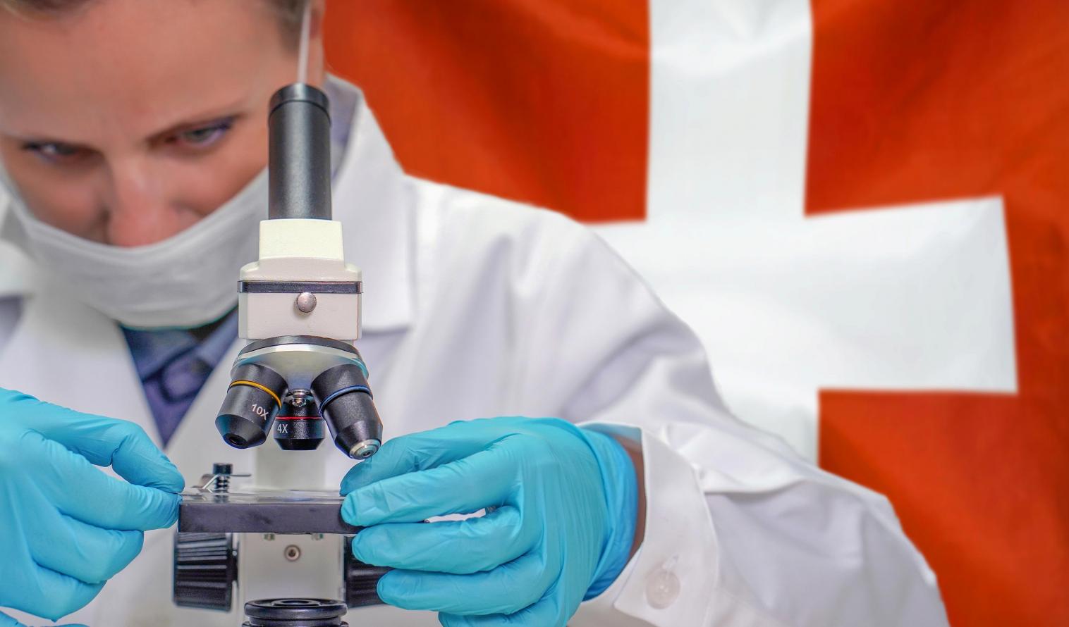 Weibliche Biochemikerin schaut durch ein Mikroskop, Schweizer Flagge im Hintergrund. 