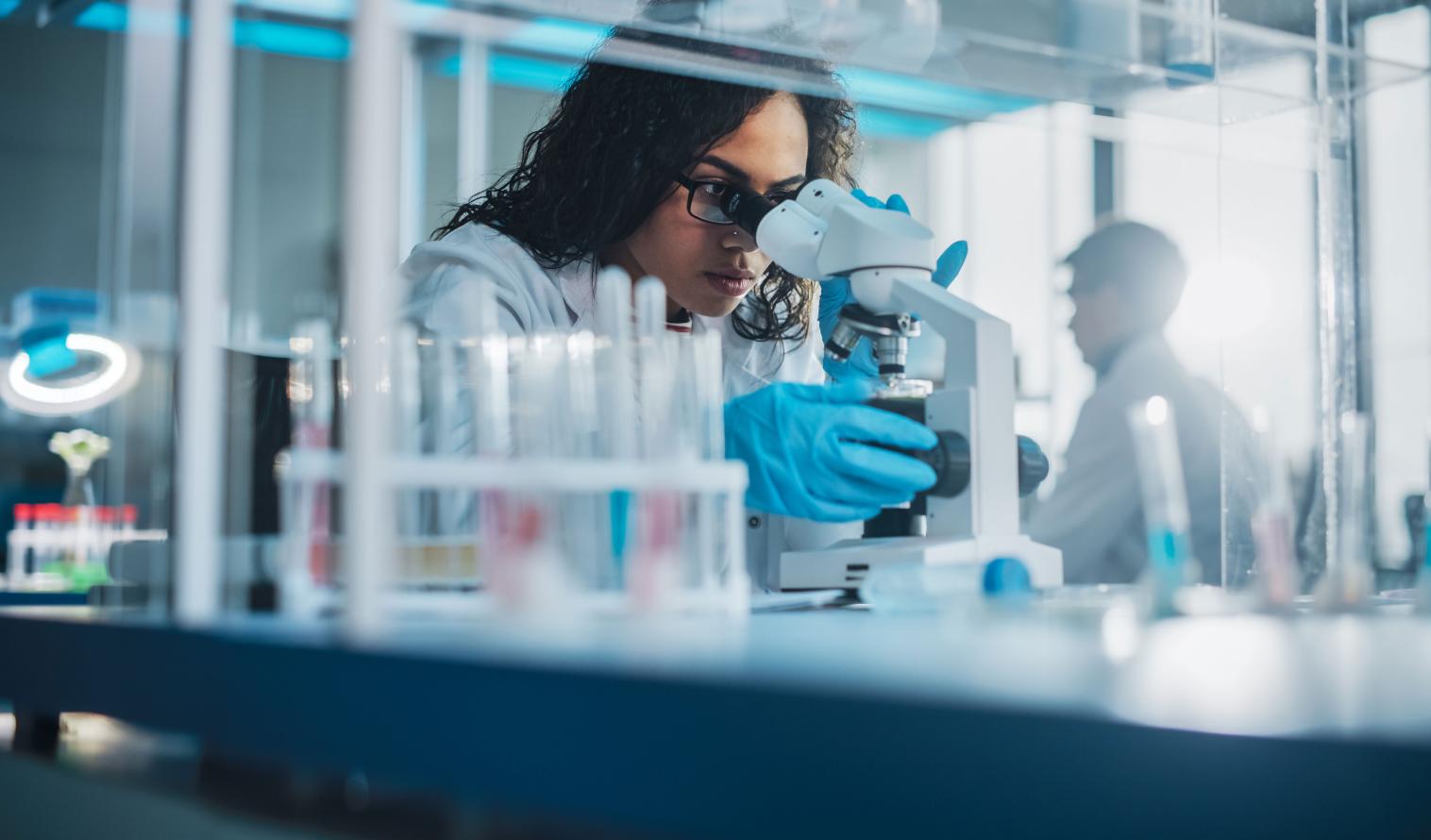 Eine Wissenschaftlerin sitzt in einem Labor und analysiert eine Testprobe unter einem Mikroskop