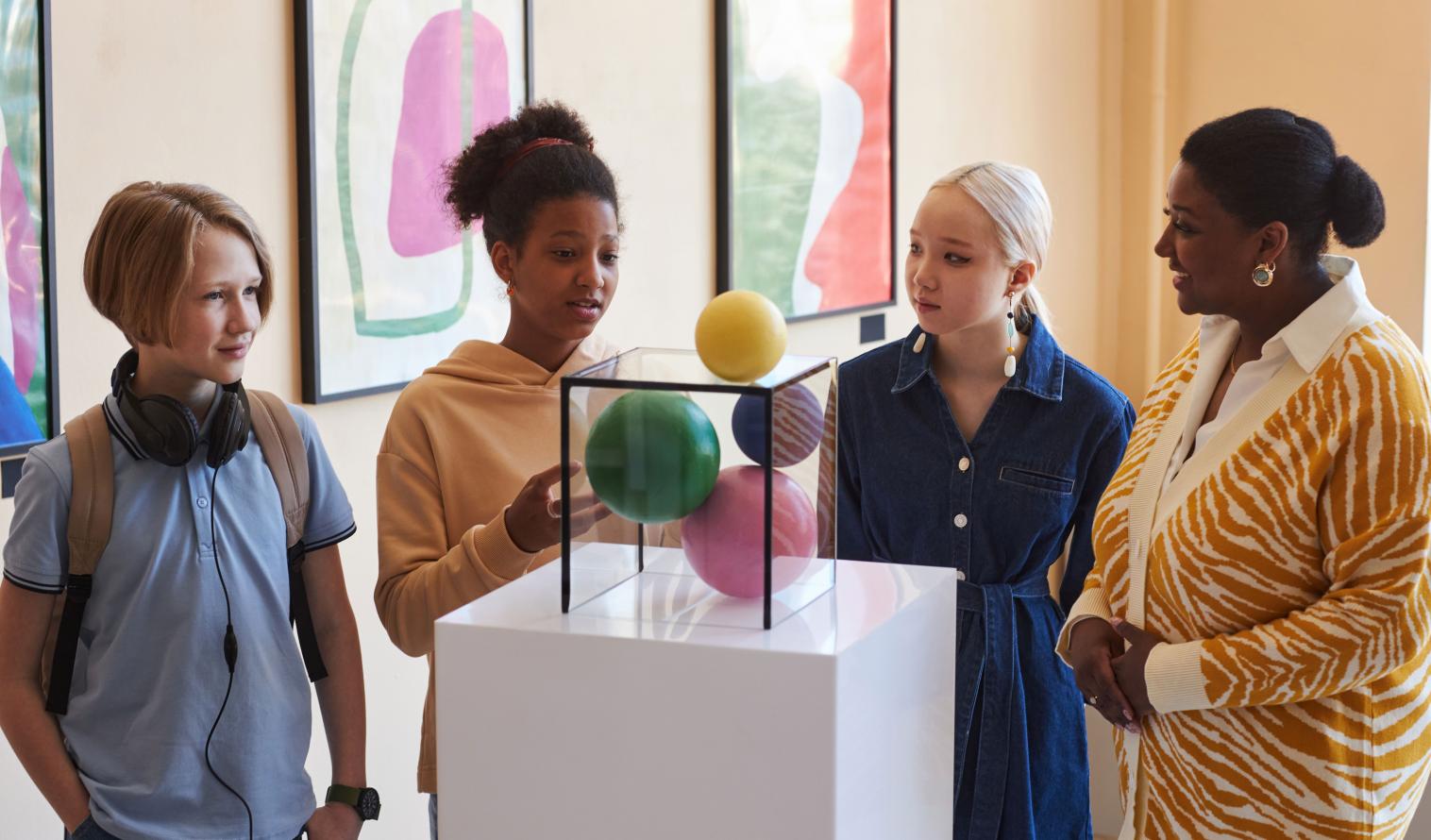 Eine vielfältige Gruppe von Teenagern steht mit einer Lehrerin bei einer Ausstellung vor einem mit bunten Bällen gefüllten Glaskasten.