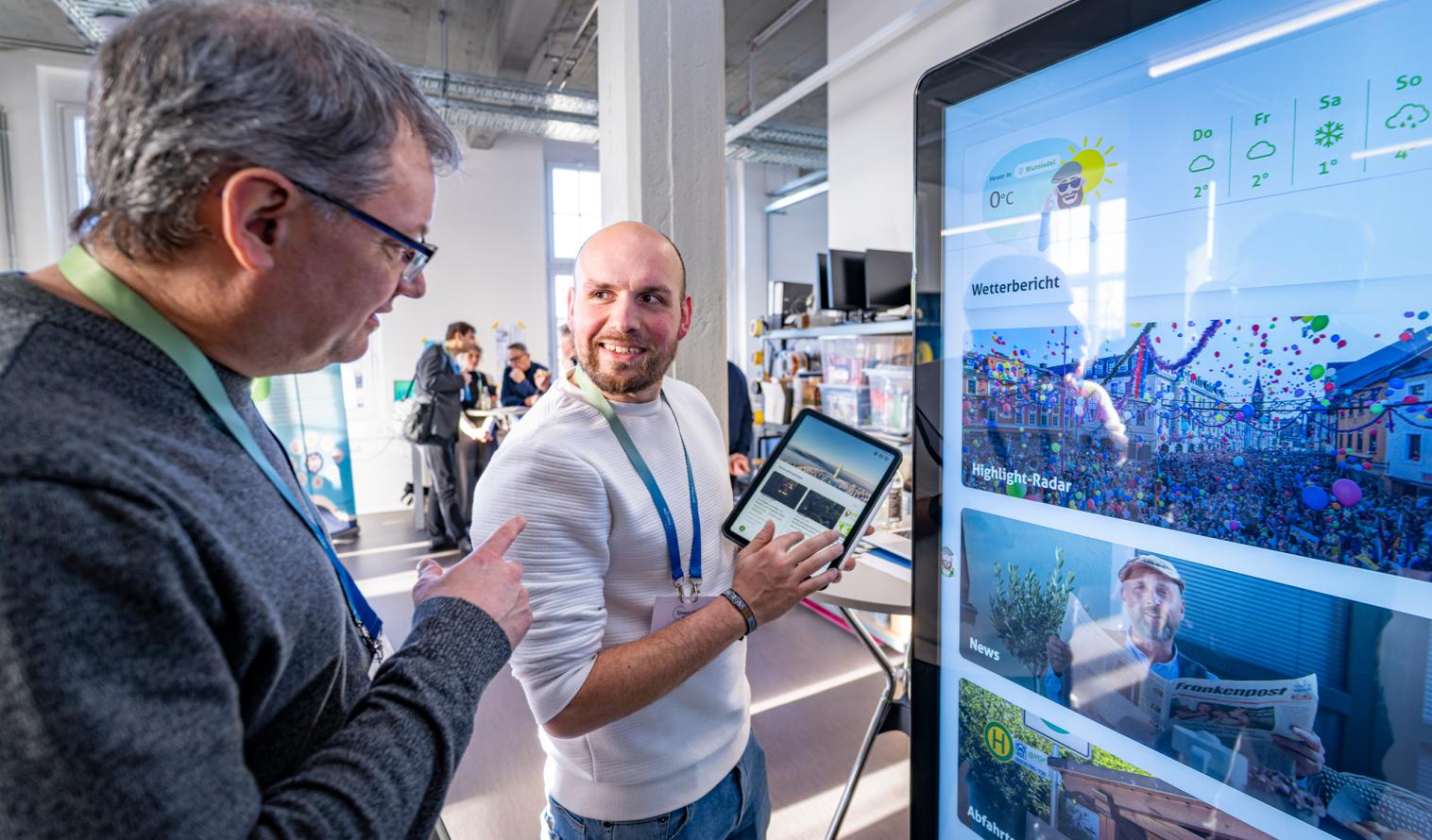 Zwei Personen stehen bei einer Ausstellung der Regionalkonferenz vor einem Bildschirm mit Nachrichten