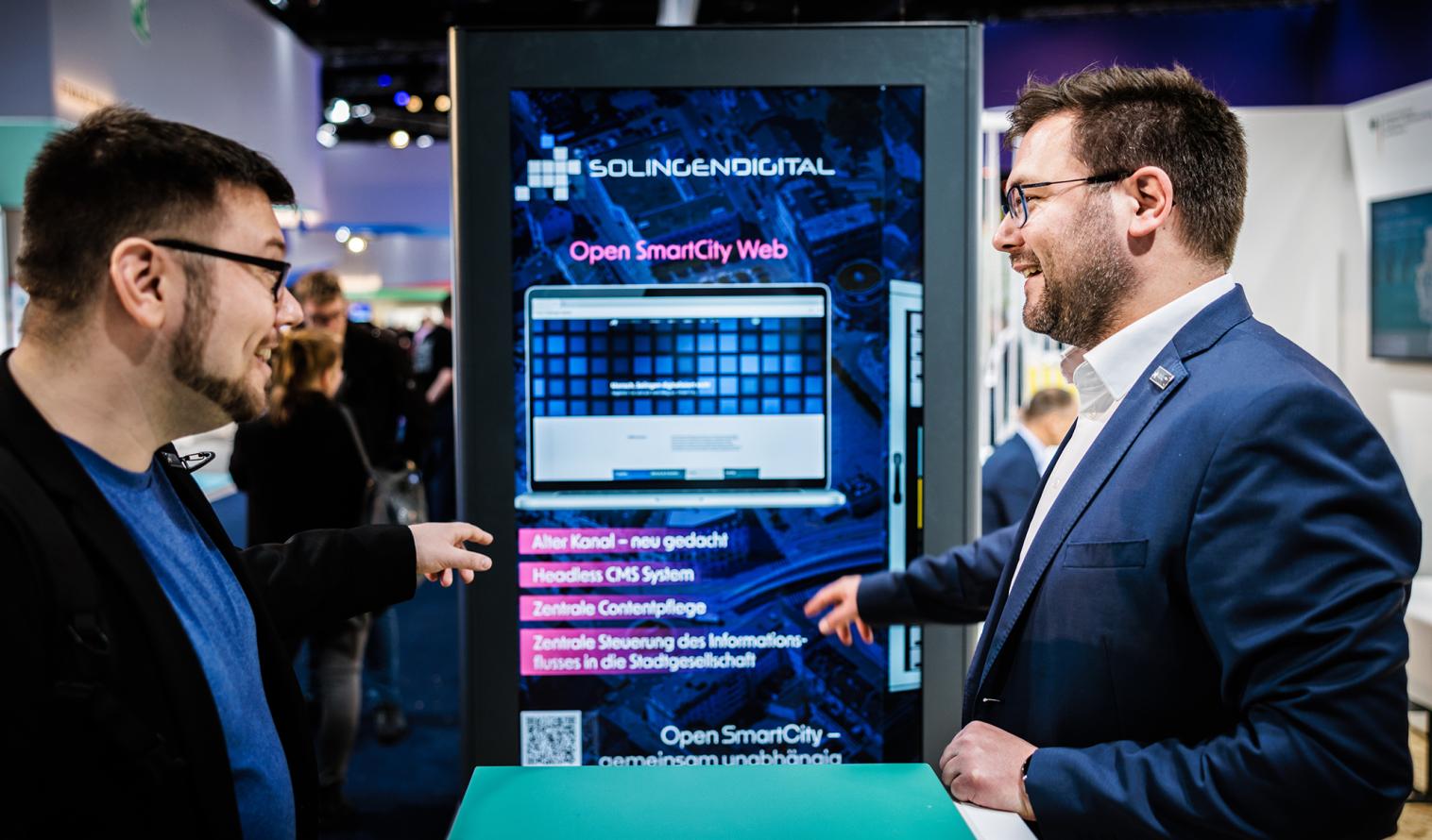 Zwei Personen auf einem Messestand vor einer digitalen Stele