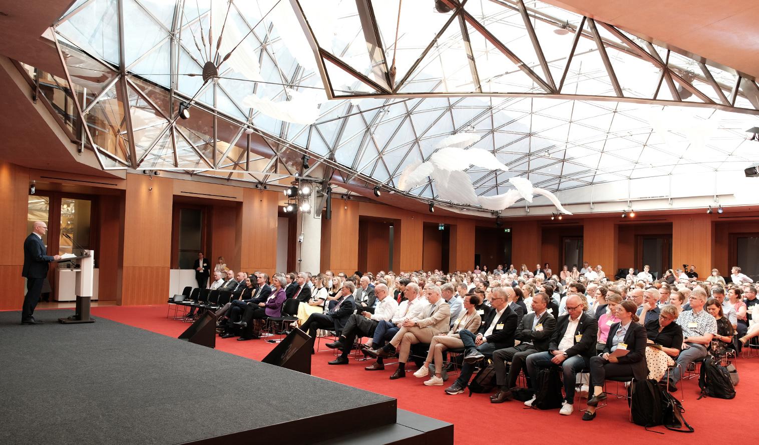 Publikum sitzt in einem großen Saal mit rotem Teppich und Glasdach und blickt auf eine Bühne