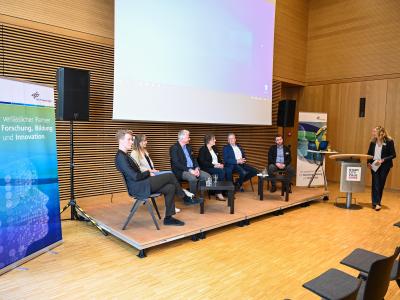 Sechs Personen sitzen mit Stühlen auf einem Podium, die Moderatorin steht an einem Tisch daneben